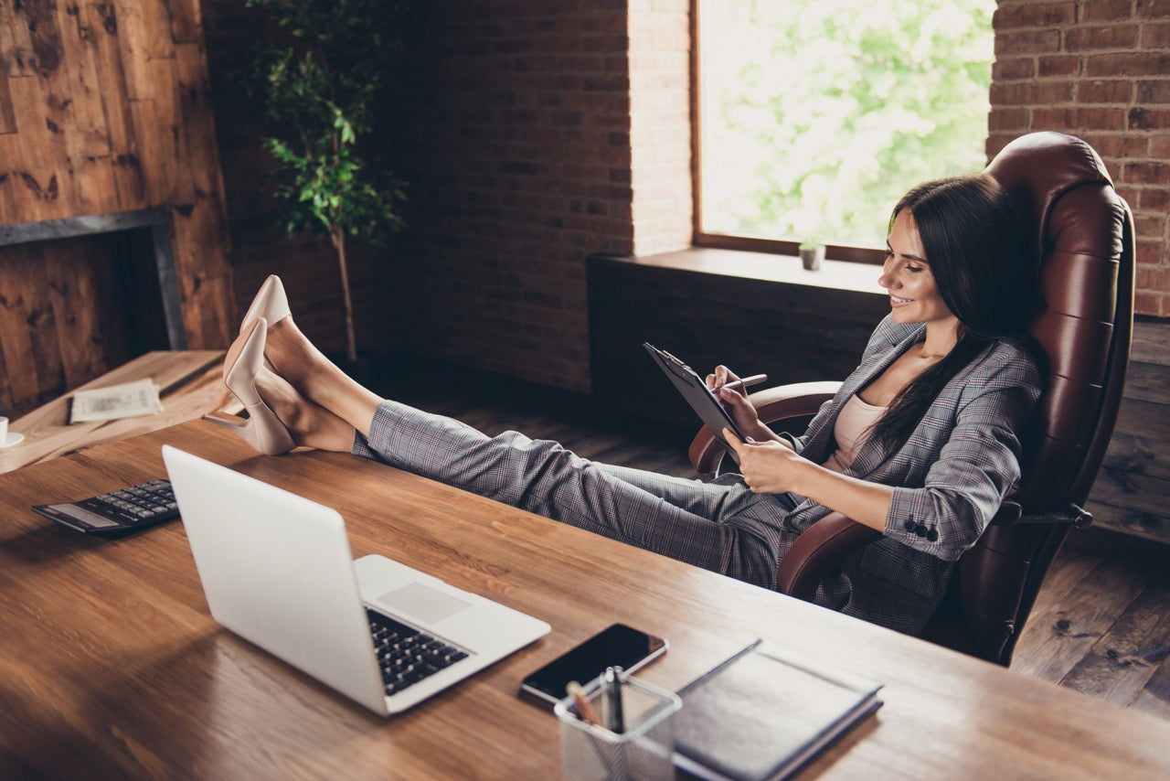 A professional woman sitting in a chair with feet up on a desk, using a tablet in a cozy office setting wearing a mid-compression bodysuit underneath. Alignelle - French-Founded